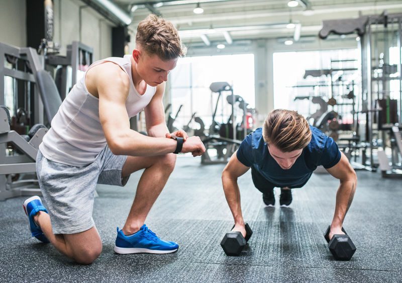Young Fit Men In Gym Doing Push Ups On Dumbbells, Measuring Time On Smartwatch.